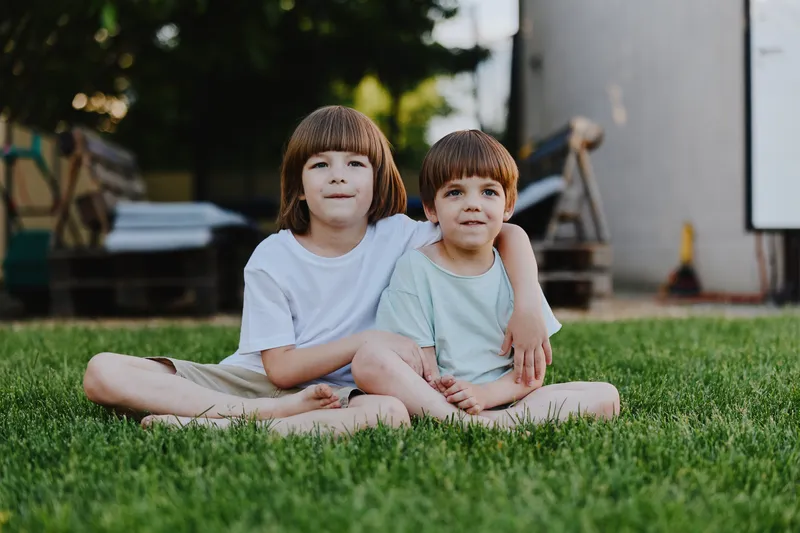 Two young boys sitting together on lush green grass, enjoying a sunny day with cheerful expressions and relaxed poses, capturing the essence of childhood friendship.