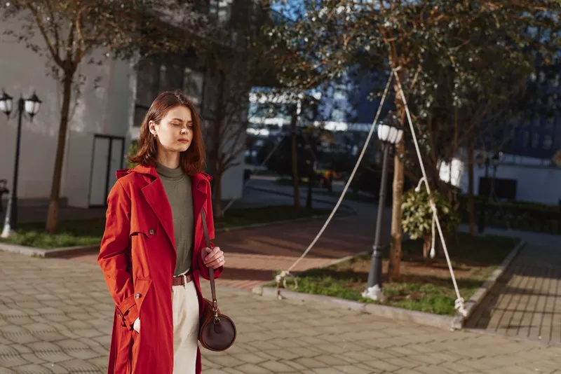 A stylish woman in a red trench coat stands on an urban street, clutching a small bag, showcasing confident street fashion on a sunny day with modern buildings in the background.