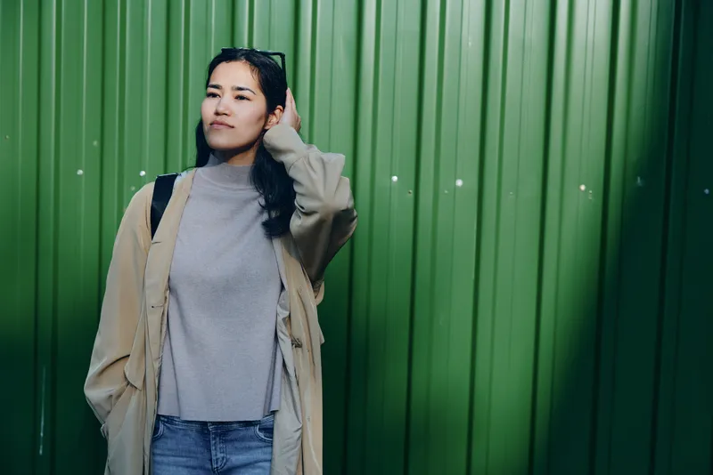 Woman in gray sweater and beige coat leans against a green metal wall, casual street fashion shot, moody daylight, calm pose, urban backdrop, wardrobe style, everyday wear, minimal accessories