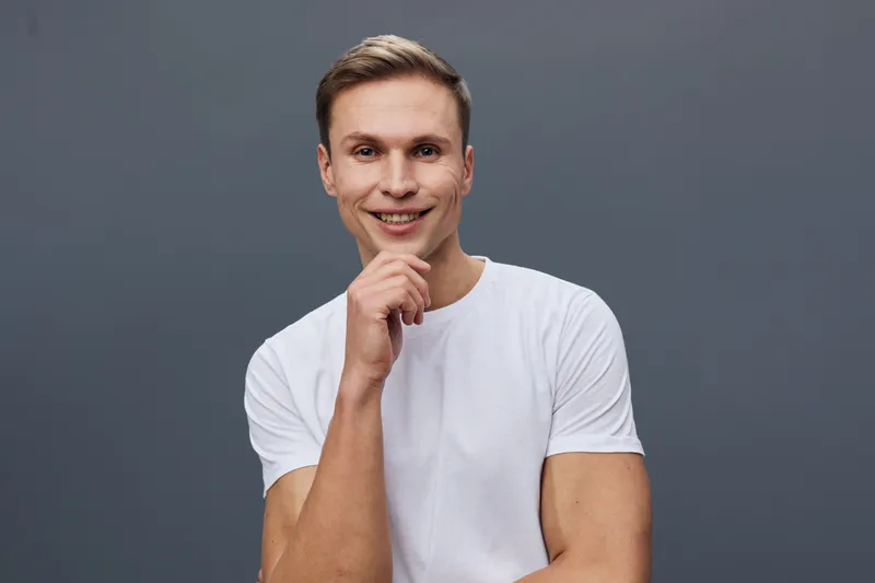 Smiling man young adult caucasian with short hair wearing casual white t-shirt looking at camera pose confident relaxed Studio portrait isolated gray background People lifestyle concept