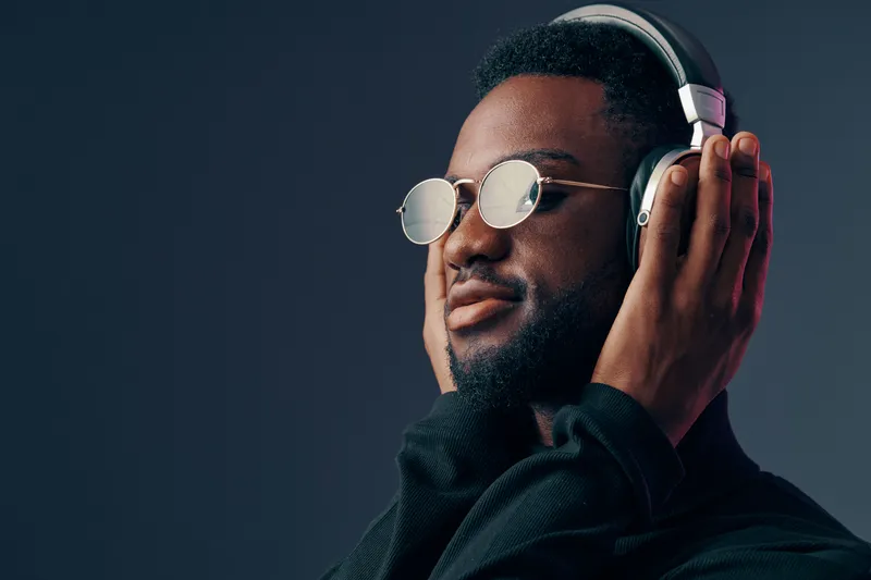 Young Black man enjoying music with headphones on, wearing stylish sunglasses, set against a dark gradient background showcasing a modern and vibrant vibe