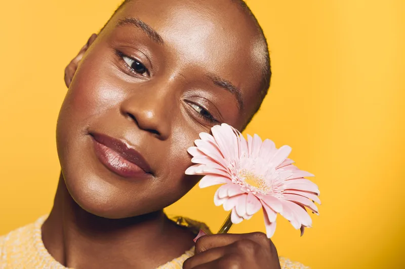Close-up of a happy African woman with dark skin holding a pink flower near her face on yellow background. Concept of natural beauty, skincare, wellness, and floral aesthetics.