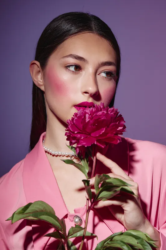 Portrait of a young woman with bright pink makeup and purple lipstick holding a vivid pink flower. She wears a matching pink suit jacket and pearl necklace in a studio setting.