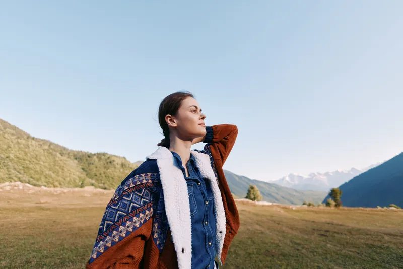 Woman portrait in mountains and nature, outdoor scene with cozy sweater standing in meadow, looking into distance. Scenic travel lifestyle, calm expression and warm autumn light.