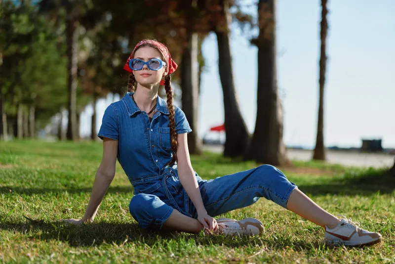Lifestyle woman in modern boho-western denim street style sitting on grass outdoors, wearing sunglasses and red bandana, enjoying sunny day with relaxed expression, film color.