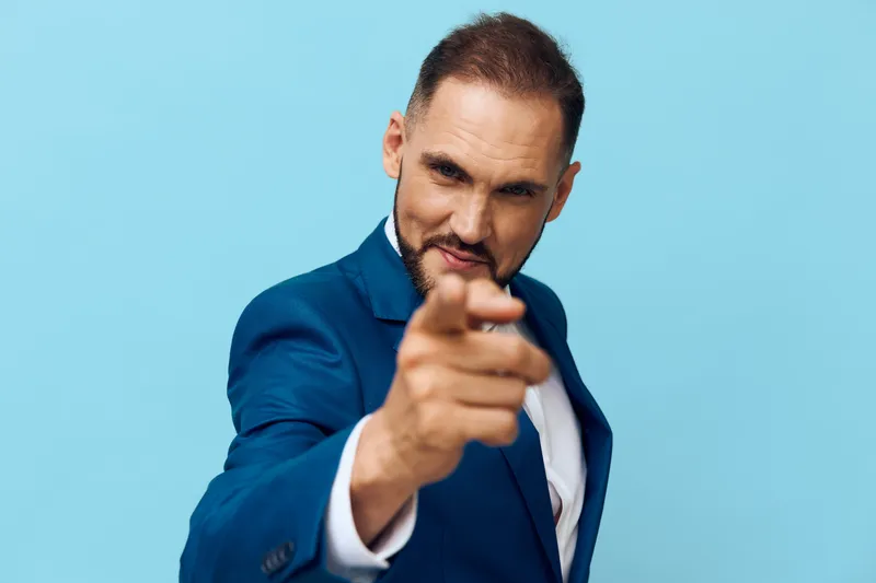 Professional man in business attire stands against a solid colored background, exuding confident energy and focus. A strong pose and direct gesture convey leadership, commitment, and clear