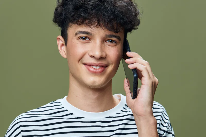 smiling young man wearing striped shirt talking on smartphone with cheerful expression against olive green background