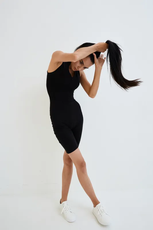 Woman tying long hair wearing black sleeveless jumpsuit and white sneakers, standing on white background, capturing casual and minimalistic sporty look.