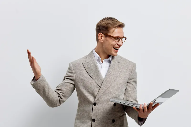 Professional smiling man in beige blazer holds open laptop while gesturing with one hand against plain white background. Business casual, technology, communication concept.
