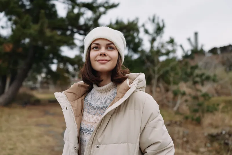 Woman in a warm beige coat and knit sweater standing outdoors among trees, presenting a cozy winter portrait and calm nature mood with soft light and breeze