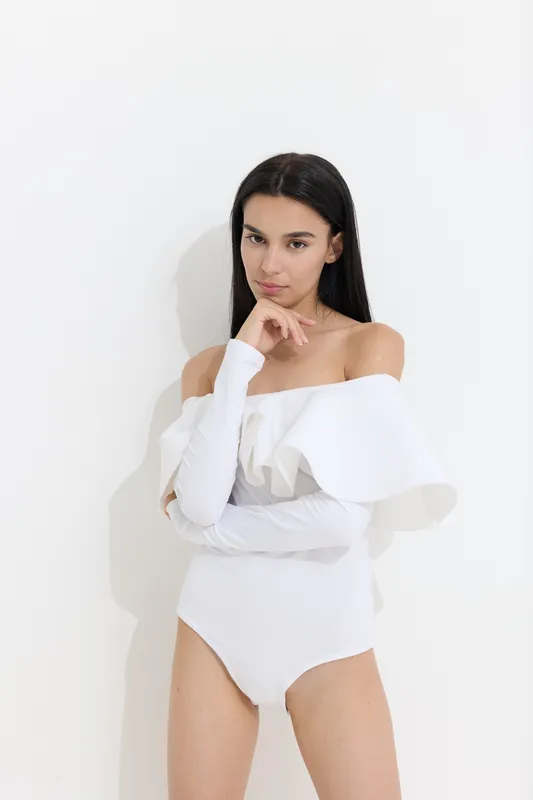 woman sport fitness portrait in white bodysuit with ruffles, long dark hair, posing with thoughtful expression against plain white background, indoor studio lighting