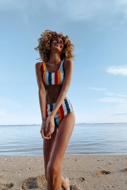 woman on beach by ocean, striped swimsuit and curly hair, standing and looking upward in sunlight, holding a towel at the waist, relaxed summer vacation concept, calm waves and blue sky