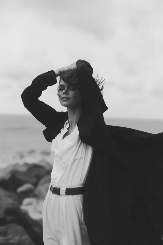 Wind sweeps a cape as a stylish woman stands on a rocky coastline, capturing monochrome fashion mood, dramatic expression, seascape and windblown hair in a timeless editorial