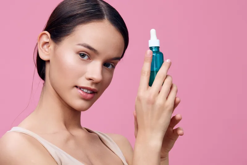 Young woman holding serum bottle on pink background, promoting skincare routine for healthy skin and hydration