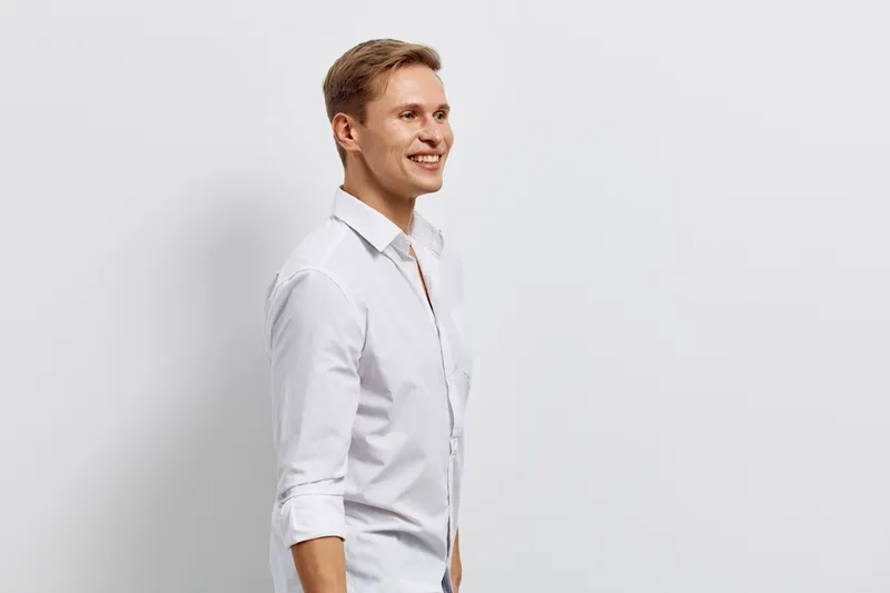 Confident young man in white shirt posing against plain white background. Studio portrait with cheerful expression, casual look, natural lighting, clean style, and minimalistic composition suitable fo