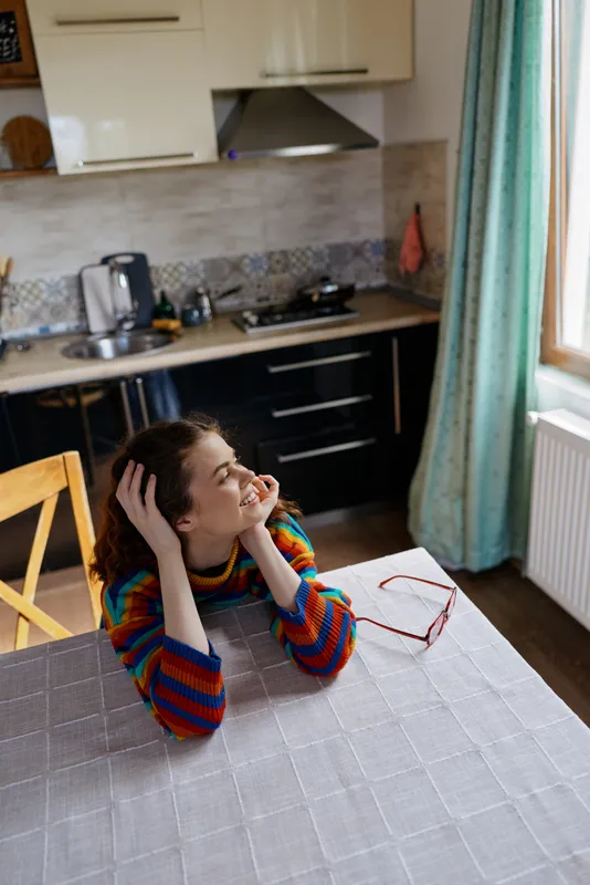 A young woman sits at a sunlit kitchen table in a striped colorful sweater, tilting her head with hands in her hair and a soft smile, capturing a relaxed home moment.