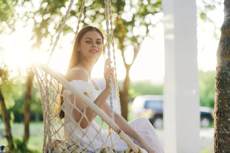 Young woman in a white dress sitting on a swing, surrounded by greenery, exuding a serene and joyful emotion during the golden hour of sunset