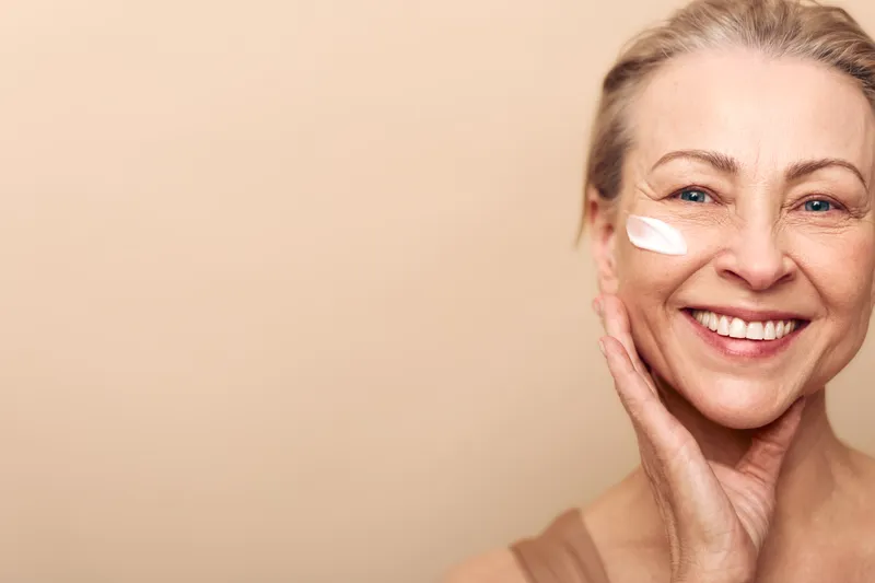 Smiling mature woman with clear skin applies moisturizing cream on cheek. Close-up portrait with natural makeup and soft lighting on beige background. Concept of skincare and antiaging beauty.