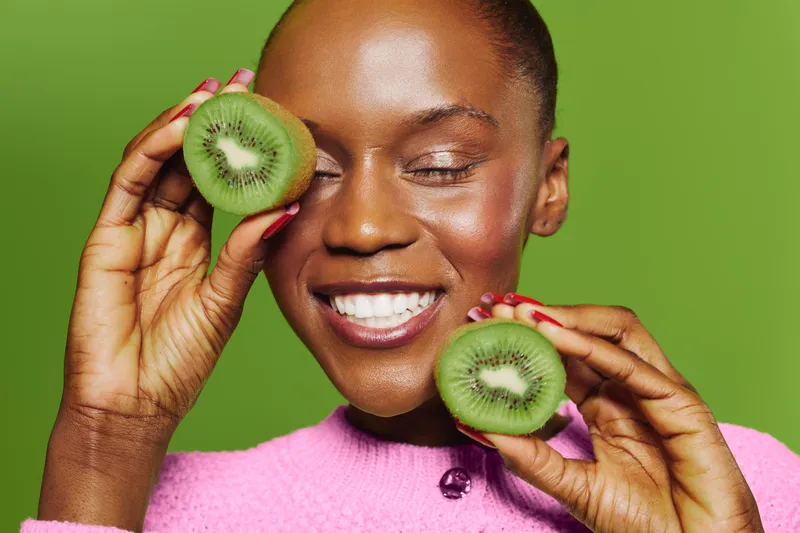 Smiling black woman with dark skin holds two halves of fresh kiwi fruit near her face, eyes closed. Bright green background and pink sweater create vibrant healthy lifestyle concept.