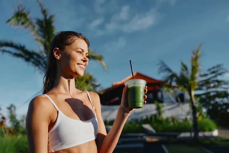 Smiling woman outdoors holding green smoothie, wearing white sports bra, sunny day, relaxed posture, tropical background, healthy lifestyle, natural light, fresh drink