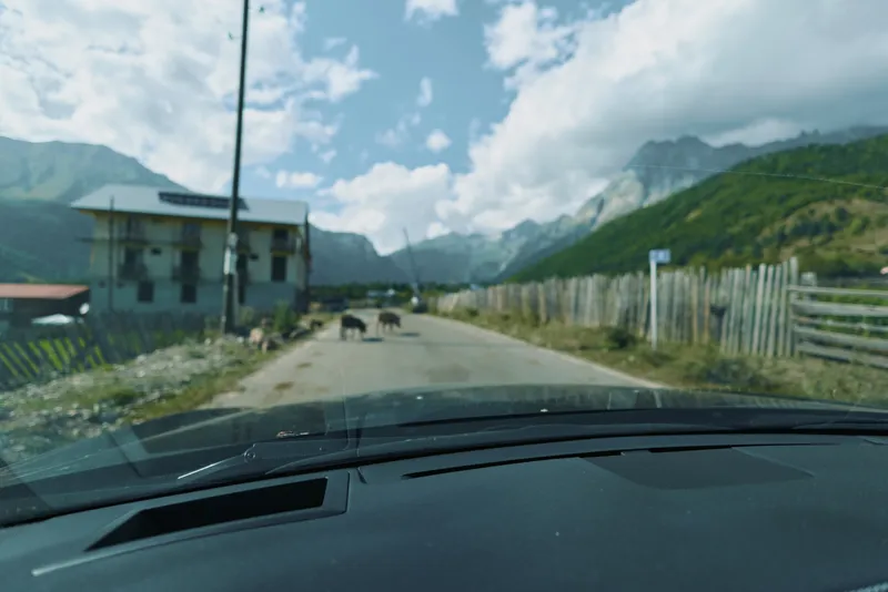 From the dashboard perspective, a driver follows a quiet rural road toward distant mountains, framed by a fence and clear sky, capturing a calm countryside drive.