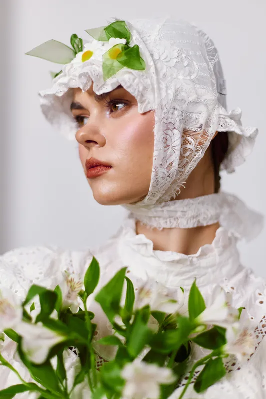 Elegant woman in a white lace dress wearing a floral headpiece, showcasing serene emotions and holding a bouquet of flowers against a soft background