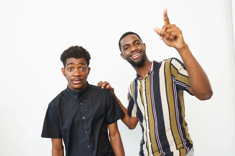 Two black men smiling together, showcasing joy and friendship in a lively atmosphere. Their attire is stylish, highlighting modern fashion trends.