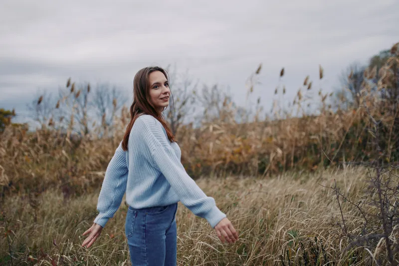 A young woman in a light blue sweater walks through windswept tall grass, captured in an authentic, natural moment that emphasizes genuine expression and believable outdoor lifestyle.