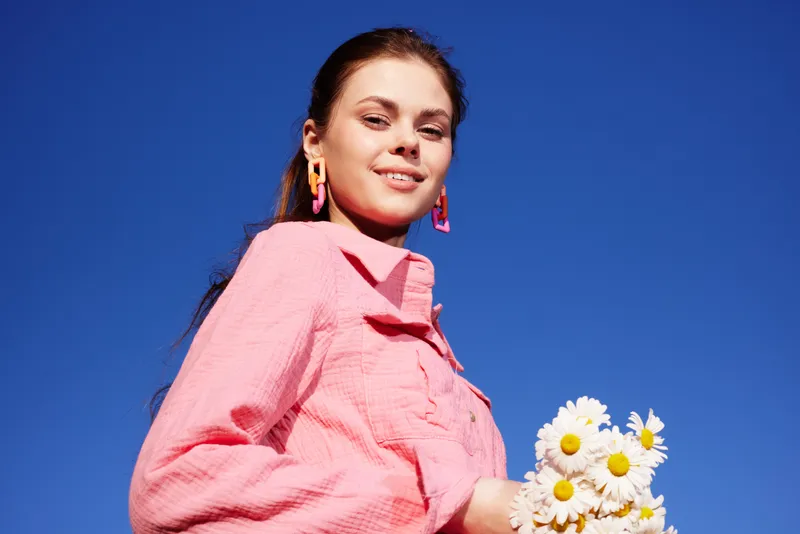 Young woman in pink shirt holding daisies against blue sky, showcasing freshness and joy, perfect for spring or summer concepts focused on nature, beauty, and happiness
