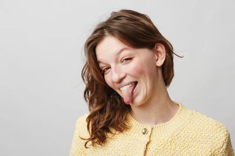 Young woman with brown hair wearing yellow sweater sticks out tongue and smiles playfully against white background. Concept of fun, humor, and casual lifestyle portrait.
