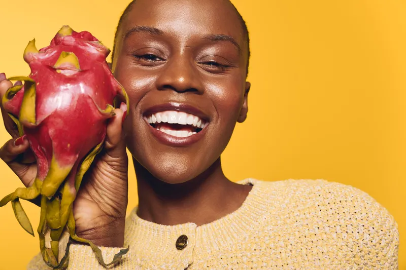Smiling dark skinned African woman holds dragon fruit close to her face against yellow background. Concept of healthy lifestyle, exotic fruit, and natural beauty with joyful expression.