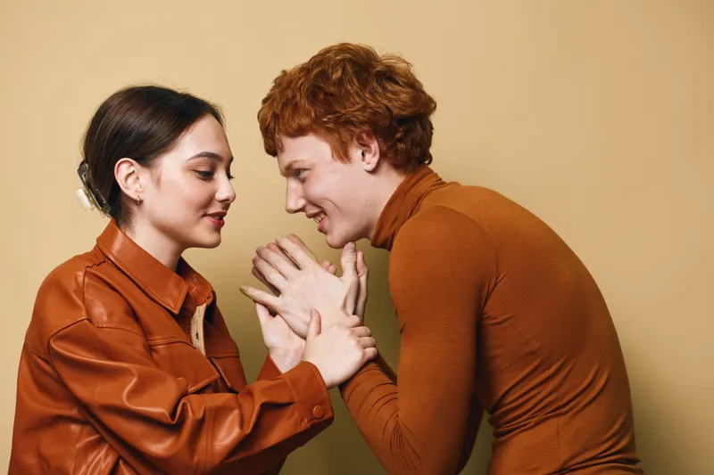 Young couple in warm clothing holding hands and smiling with closed eyes, expressing affection and connection against beige background in casual studio portrait.
