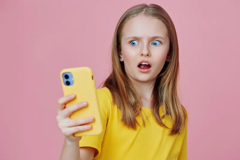 Surprised girl looking at smartphone against a pink background, wearing a yellow shirt, expressing shock and curiosity Ideal for youth related content