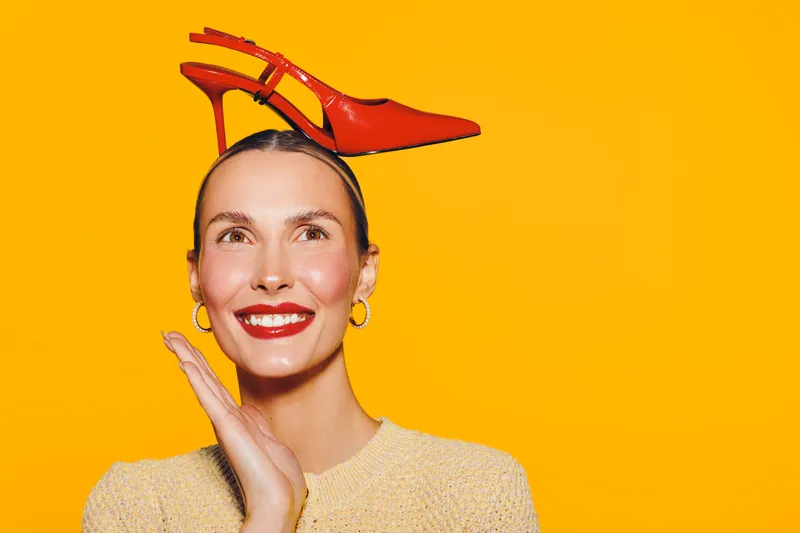 Smiling woman with red high heel shoe balanced on her head, wearing yellow sweater and gold hoop earrings. Bright yellow background creates playful and creative fashion concept.
