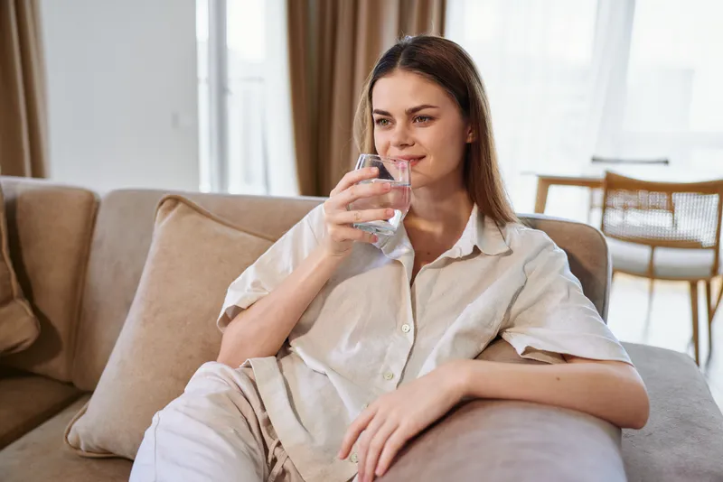 Woman drinking water from glass on sofa at home, relaxed and comfortable in casual clothes, portrait of hydration and calm in livingroom with soft natural light and cozy decor