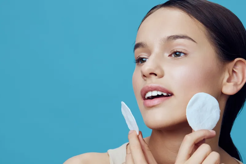 Young woman with clear skin applying skincare products, smiling and holding cotton pads against a bright blue background, showcasing a fresh and healthy beauty concept