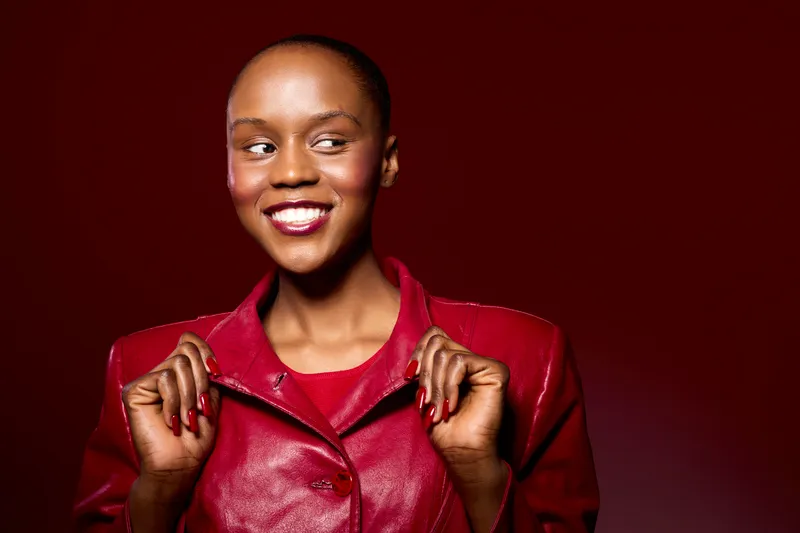 Smiling dark skinned African woman in red leather jacket looks away confidently, isolated on dark red background. Concept for fashion, beauty, confidence, and positive mood.