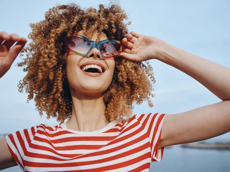 Playful portrait of a joyful woman with curly hair, wearing a red and white striped shirt and oversized sunglasses, captured outdoors under a bright sky with carefree energy.