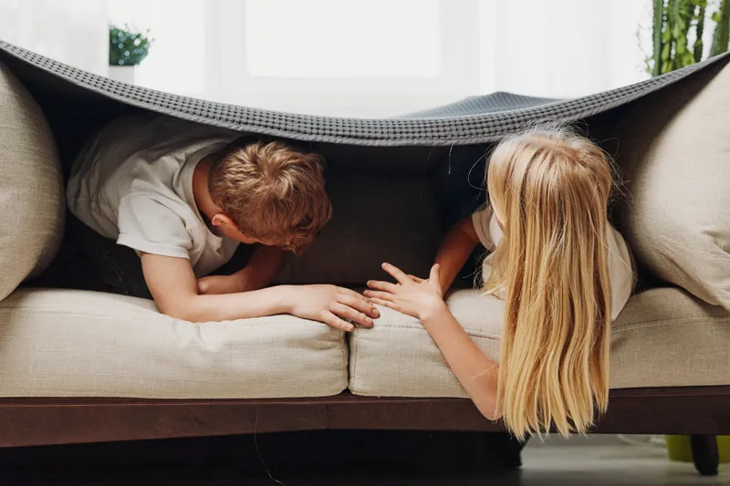 Two children play hiding under a blanket on a beige sofa in a bright room. The boy and girl interact closely, showing playful bonding and childhood imagination at home.