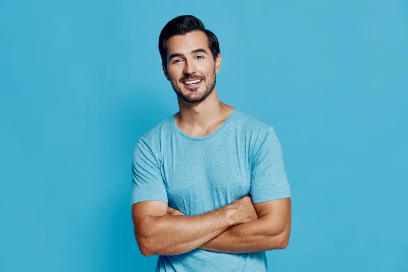 Smiling cheerful young man with crossed arms wearing a light blue shirt, looking at the camera against a bright blue background, exuding a friendly vibe.
