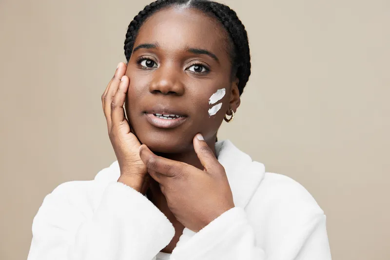 Young Black woman applying skincare on her face, promoting beauty and self care, with a calm expression and neutral background for a soothing atmosphere