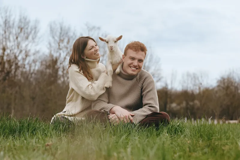 Couple outdoor lifestyle goat kid moment in a grassy meadow, smiling together among nature and warmth on a sunny day, capturing a cozy, candid family scene outdoors.