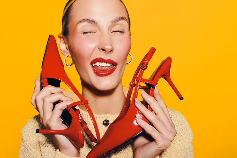 Happy woman with red lipstick holding red high heel shoes close to her face on bright yellow background. Concept of fashion, style, shopping, and joyful emotions.