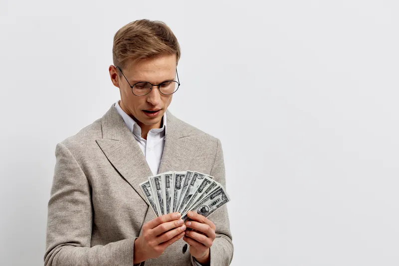 Man wearing glasses in beige blazer holding bunch of dollar bills isolated on plain light gray background studio portrait. Business and finance concept