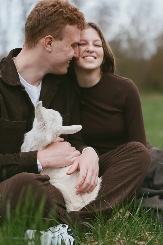 A couple shares a tender moment outdoors, cradling a small goat kid as they smile softly. Warm natural light highlights affection, companionship, and rural life.