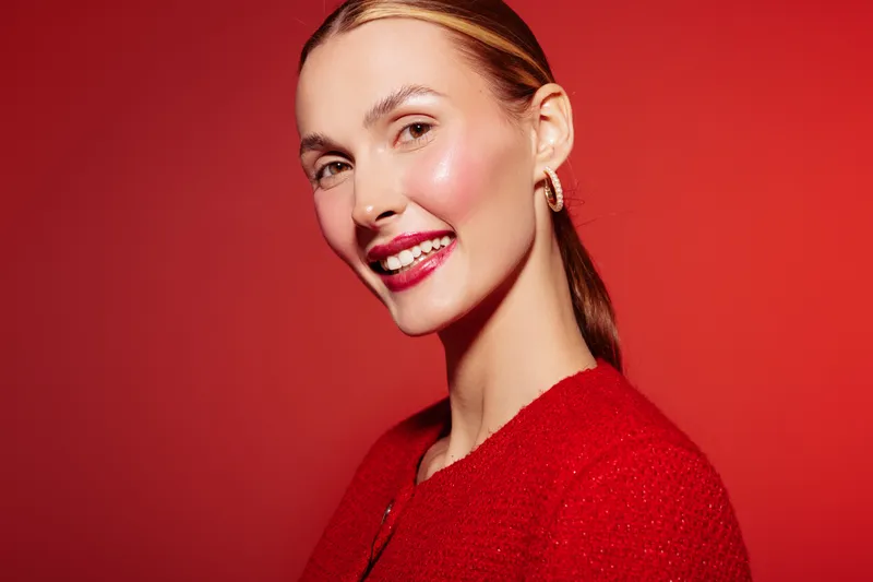 Smiling woman with shiny skin and red lipstick wears red sweater against red background. Portrait of confident female model with sleek hair and natural makeup in studio lighting.