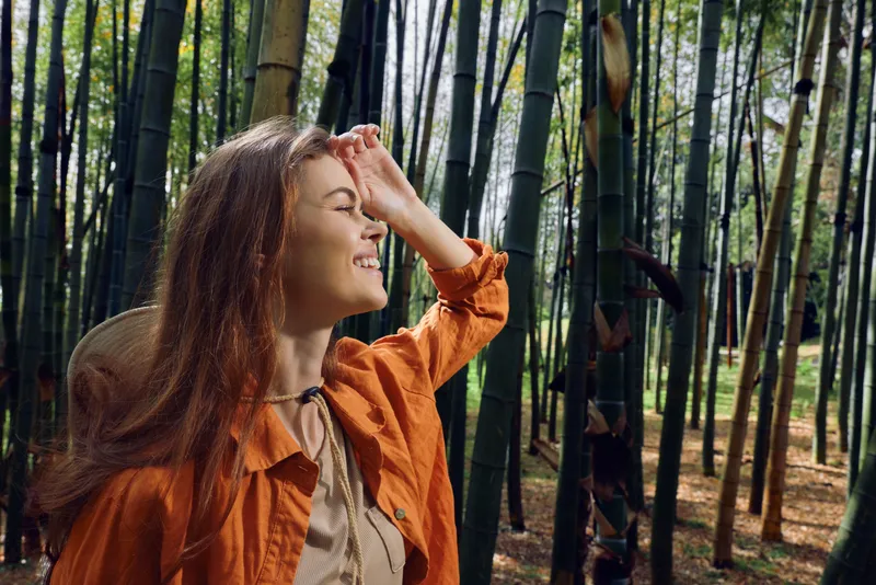 Woman portrait in a bamboo forest with a bright smile, wearing a straw hat and orange jacket, sunlight filtering through stalks, nature scene conveying joy and peaceful outdoor exploration.