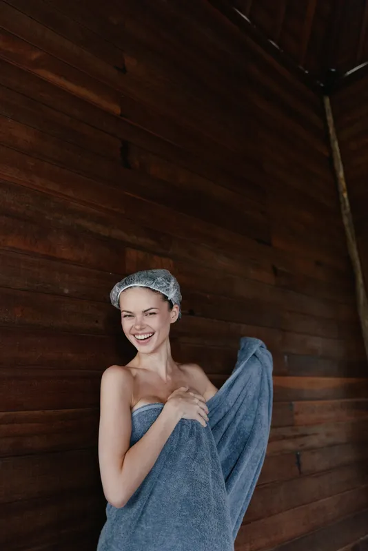 Smiling woman wrapped in blue towel with shower cap, standing by wooden wall, expressing joy and freshness in a cozy spa environment.