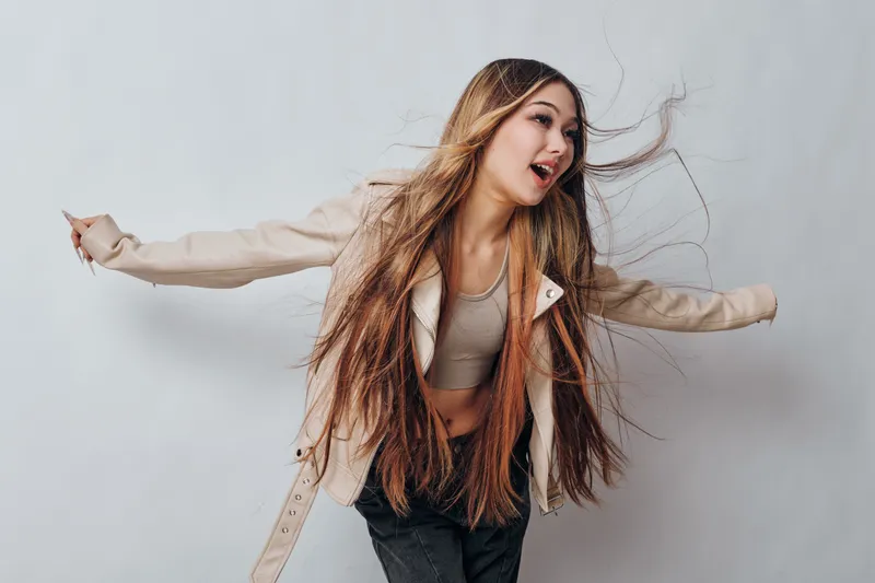 Young woman with long hair wearing casual jacket and crop top dancing or posing with arms wide open against plain neutral background. Energetic girl with happy expression and movement.