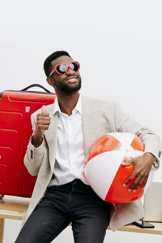 Confident man at the beach with suitcase and beach ball enjoying vacation time, dressed in casual stylish outfit, sunglasses, smiling sunny day holiday concept relaxed leisure travel.